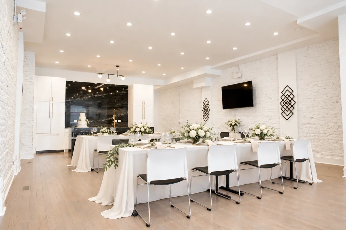 Intimate wedding dinner at 3535 Venue: long tables with ivory linens, white flowers and greenery garlands, and a wedding cake by the black marble kitchen (virtually staged)