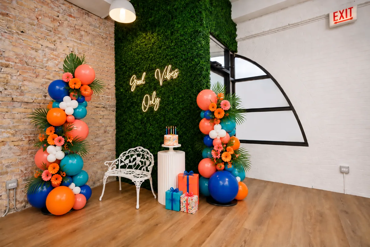 Birthday setup at Montrose Studio: coral, orange and blue balloon columns, a cake on a white pedestal and gifts in front of the “Good Vibes Only” neon green wall (virtually staged)