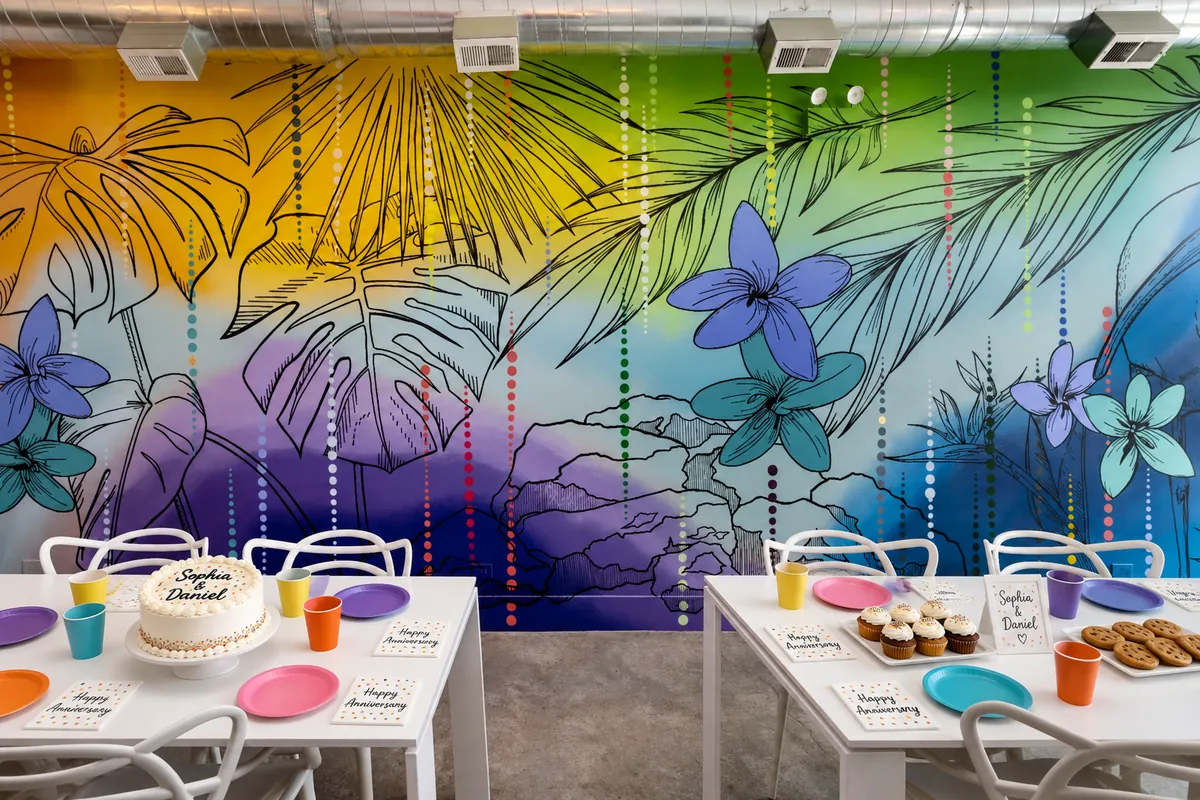 Birthday party setup at Nala’s Party Room: white tables with colorful plates and cups, a cake, cupcakes and cookies in front of the tropical mural (virtually staged)