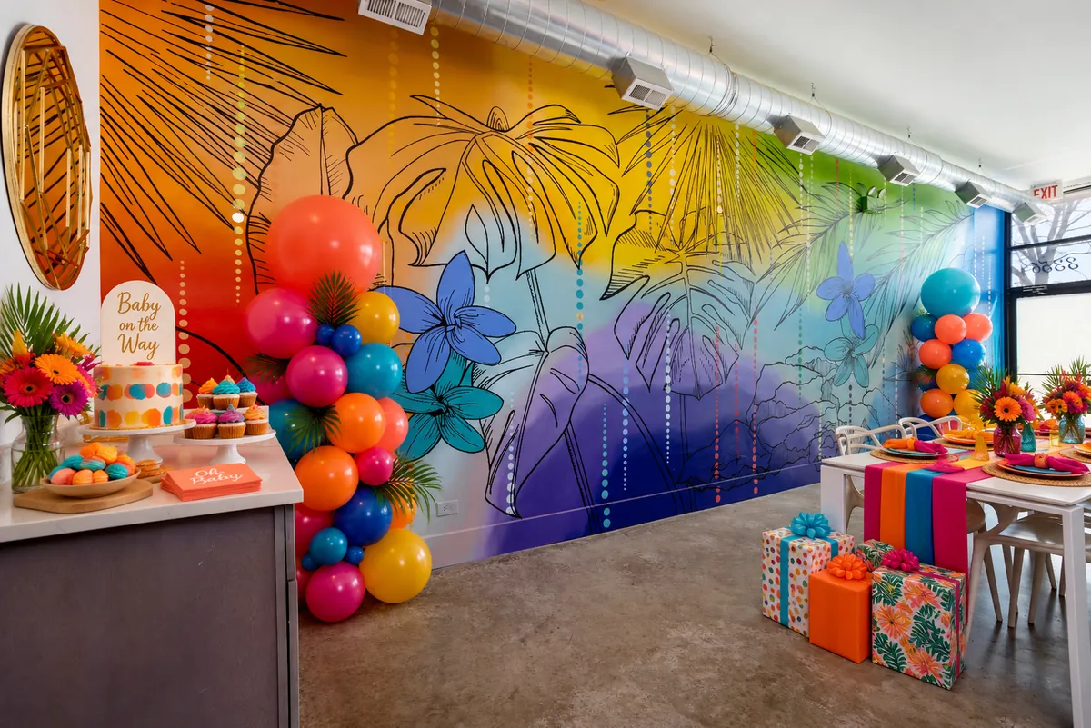 Baby shower setup at Nala’s Party Room: colorful balloon columns, a “Baby on the Way” cake, cupcakes, gifts and bright table runners in front of the tropical mural (virtually staged)