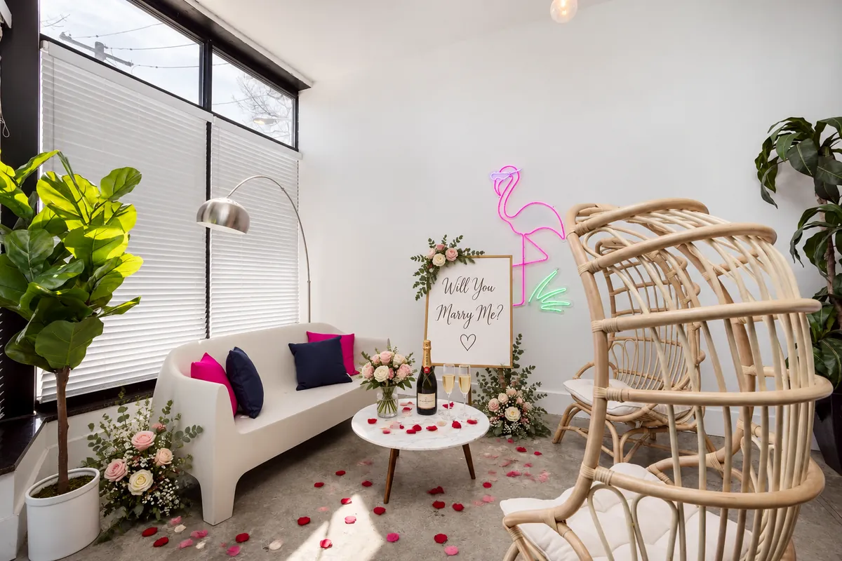 Proposal setup at Nala’s Party Room: the white lounge sofa with roses, champagne and a Will You Marry Me sign under the neon flamingo (virtually staged)
