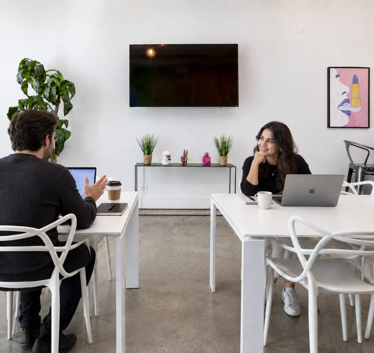 Two people working on laptops at the white tables at Nala’s Party Room, with the wall-mounted TV behind them (virtually staged)
